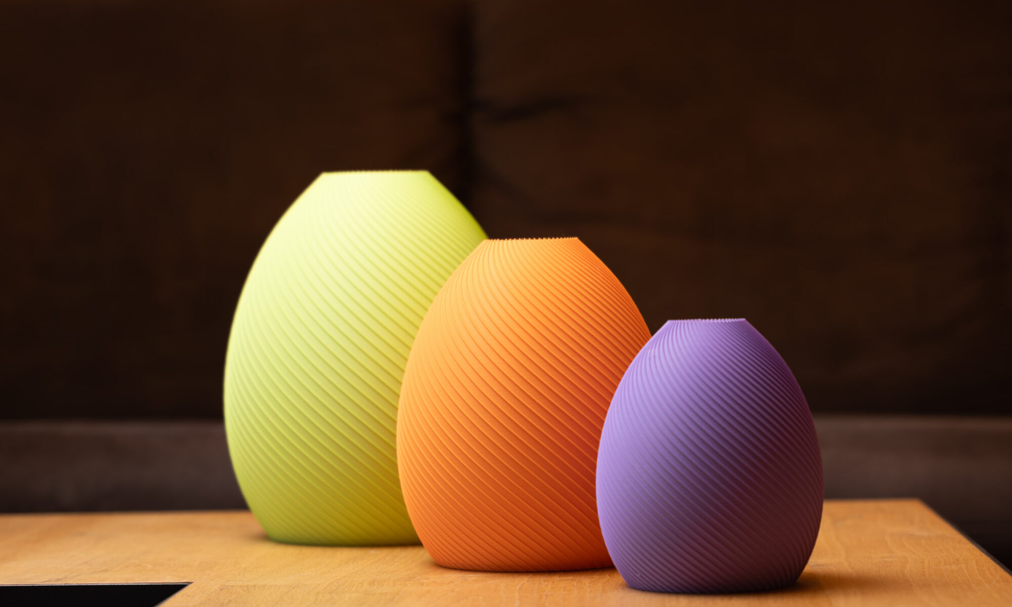 Drei elegante, farbige 3D-gedruckte Vasen in Gelb, Orange und Lila mit feiner Rillenstruktur auf Holzoberfläche.