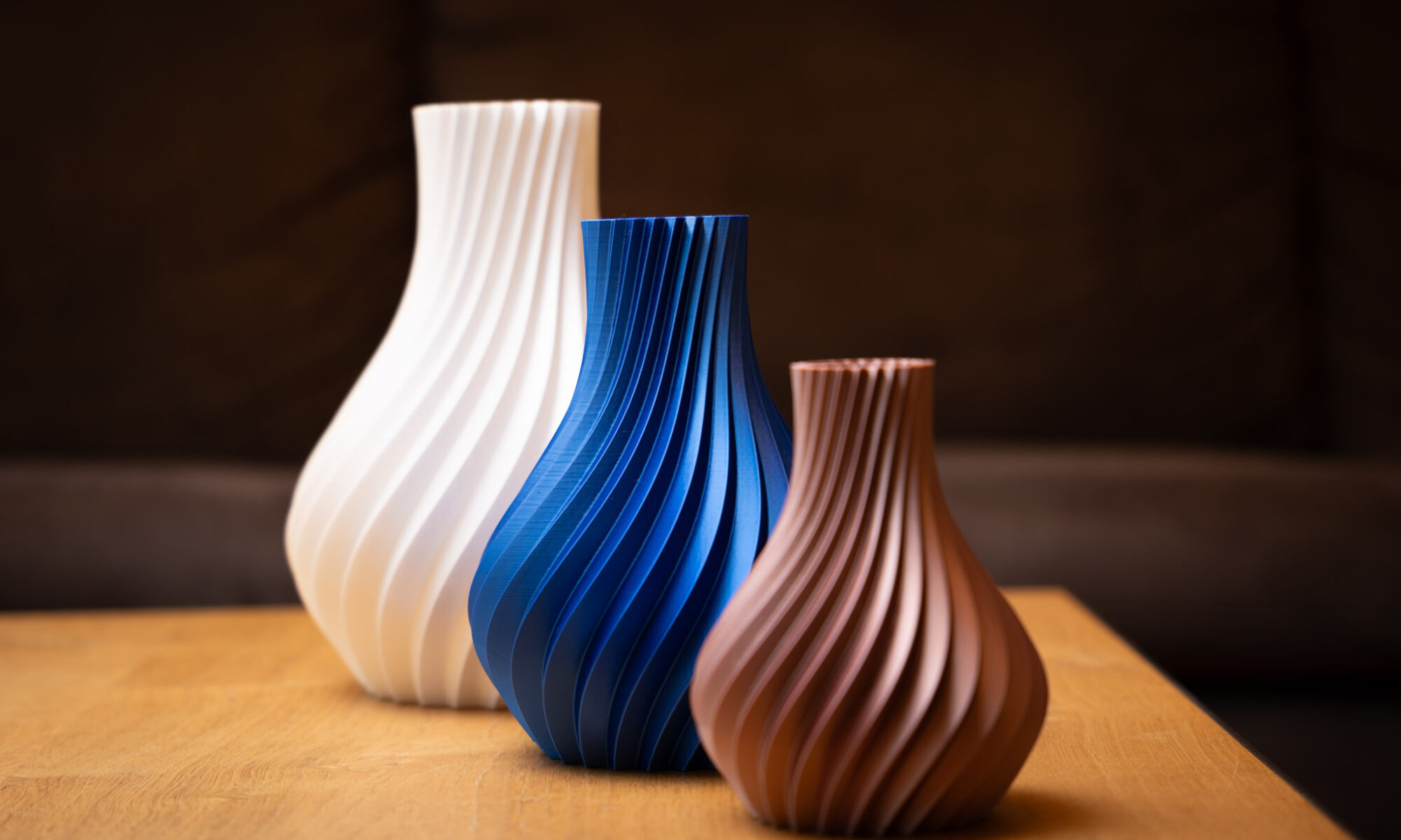 Drei elegante 3D-gedruckte Vasen in Weiß, Blau und Kupfer mit spiralförmigem Design auf einem Holztisch.