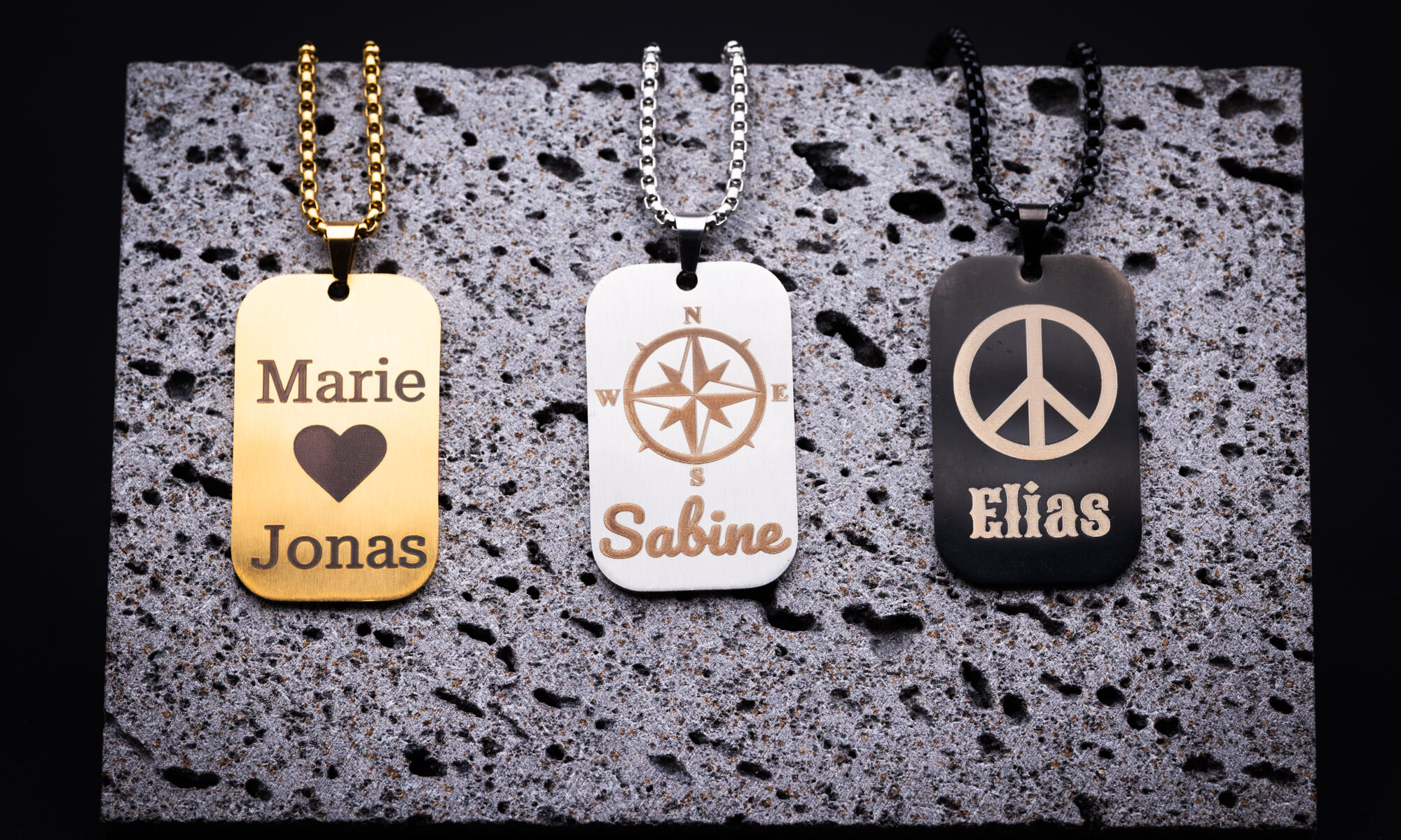 Drei stilvolle Namensanhänger aus 3D-Druck: Gold mit Herzmotiv, Silber mit Kompass, Schwarz mit Peace-Zeichen. Elegant designt.